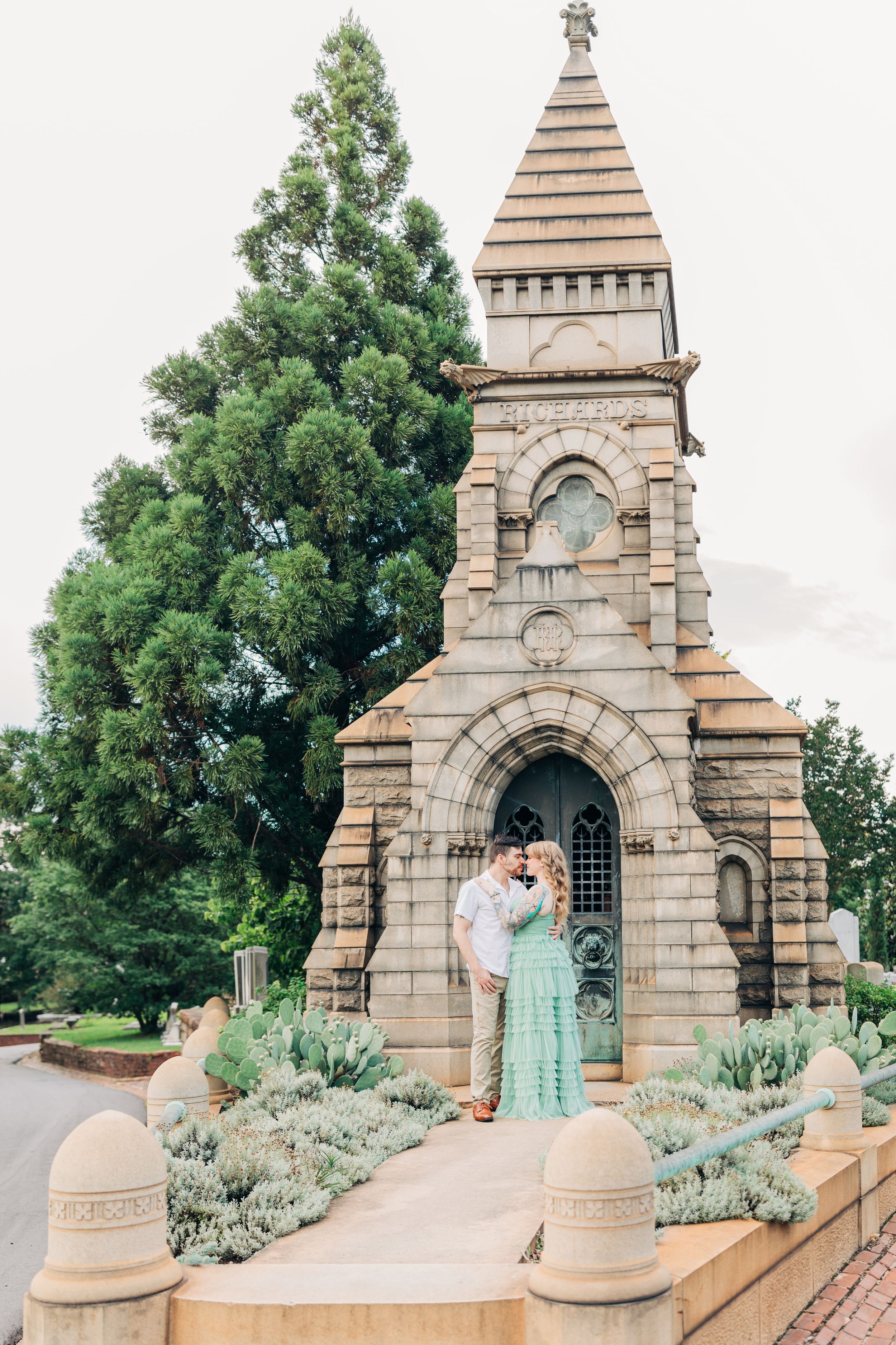 Couple standing at entrance of stone Gothic chapel with ornate spire and decorative landscaping.