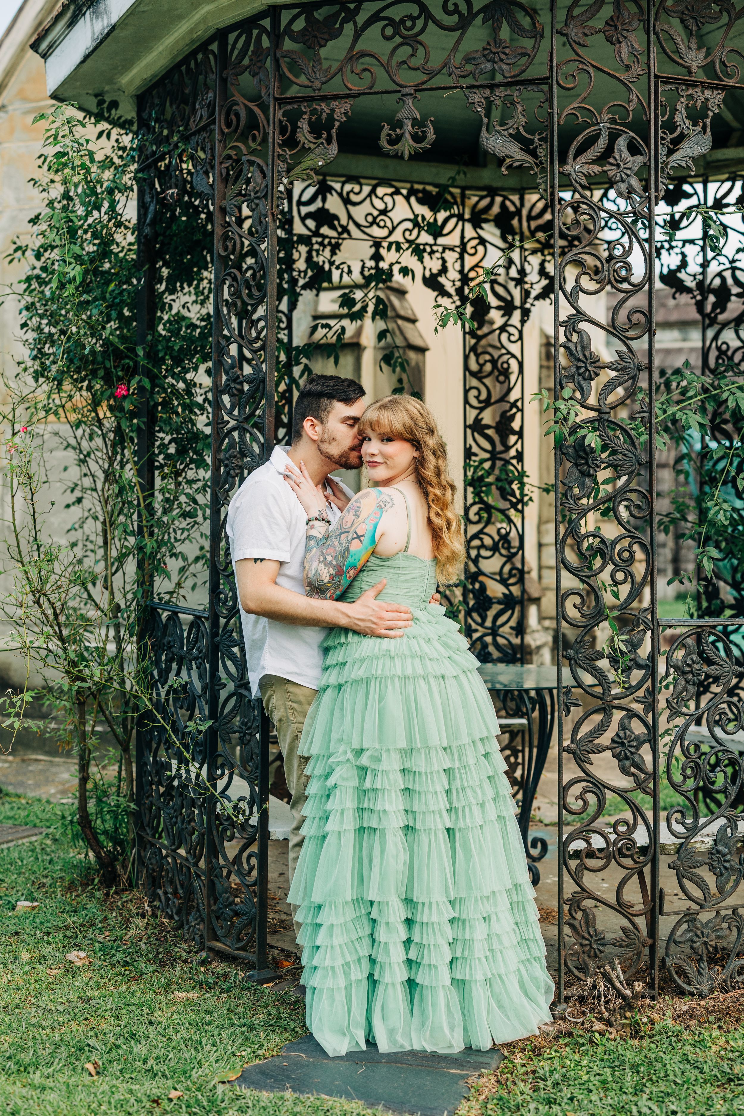 A romantic couple embraces in an ornate wrought iron gazebo, with the person wearing a flowing mint green tiered gown.