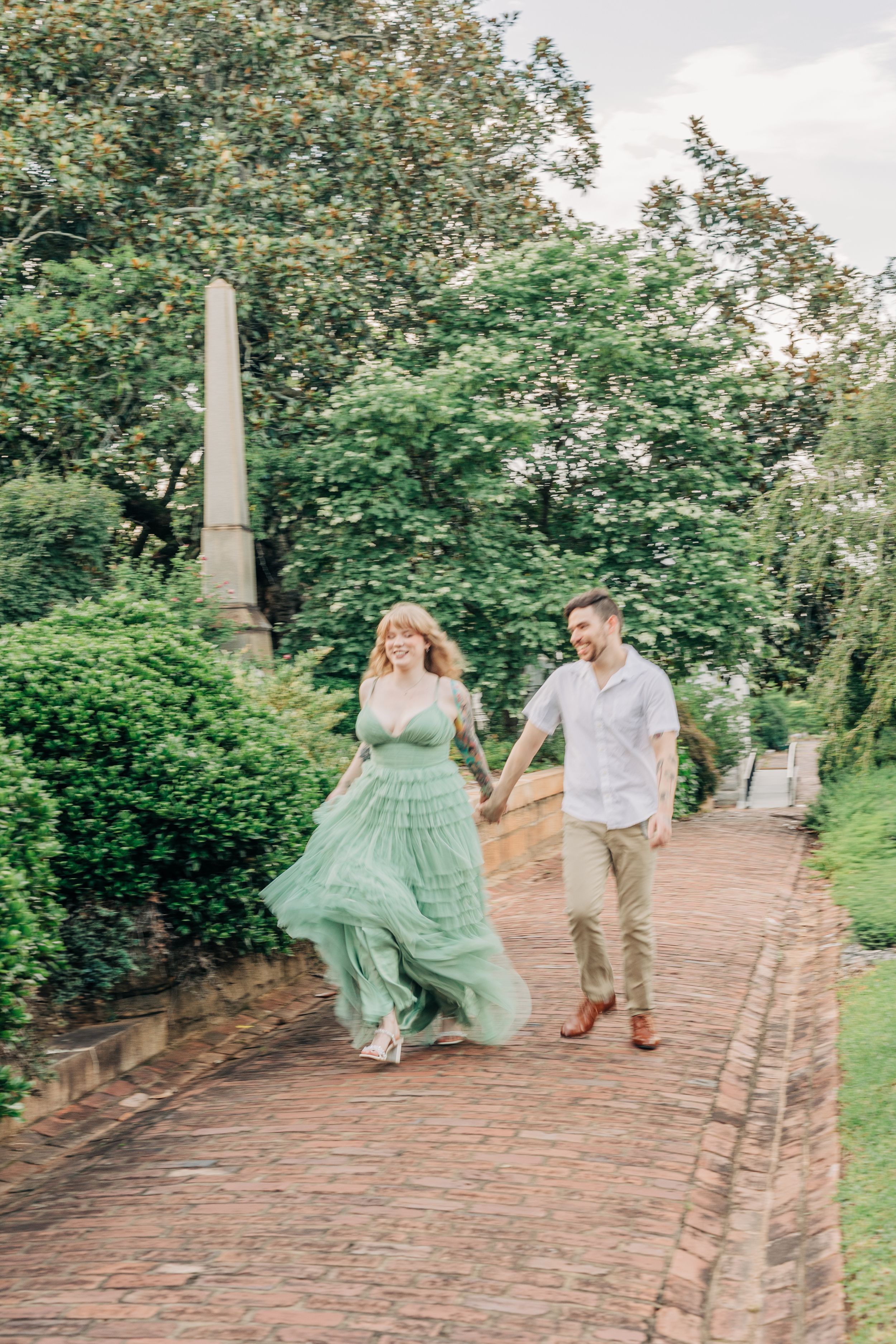 A couple strolls together on a brick path through a garden, with the person in a flowing mint green dress.