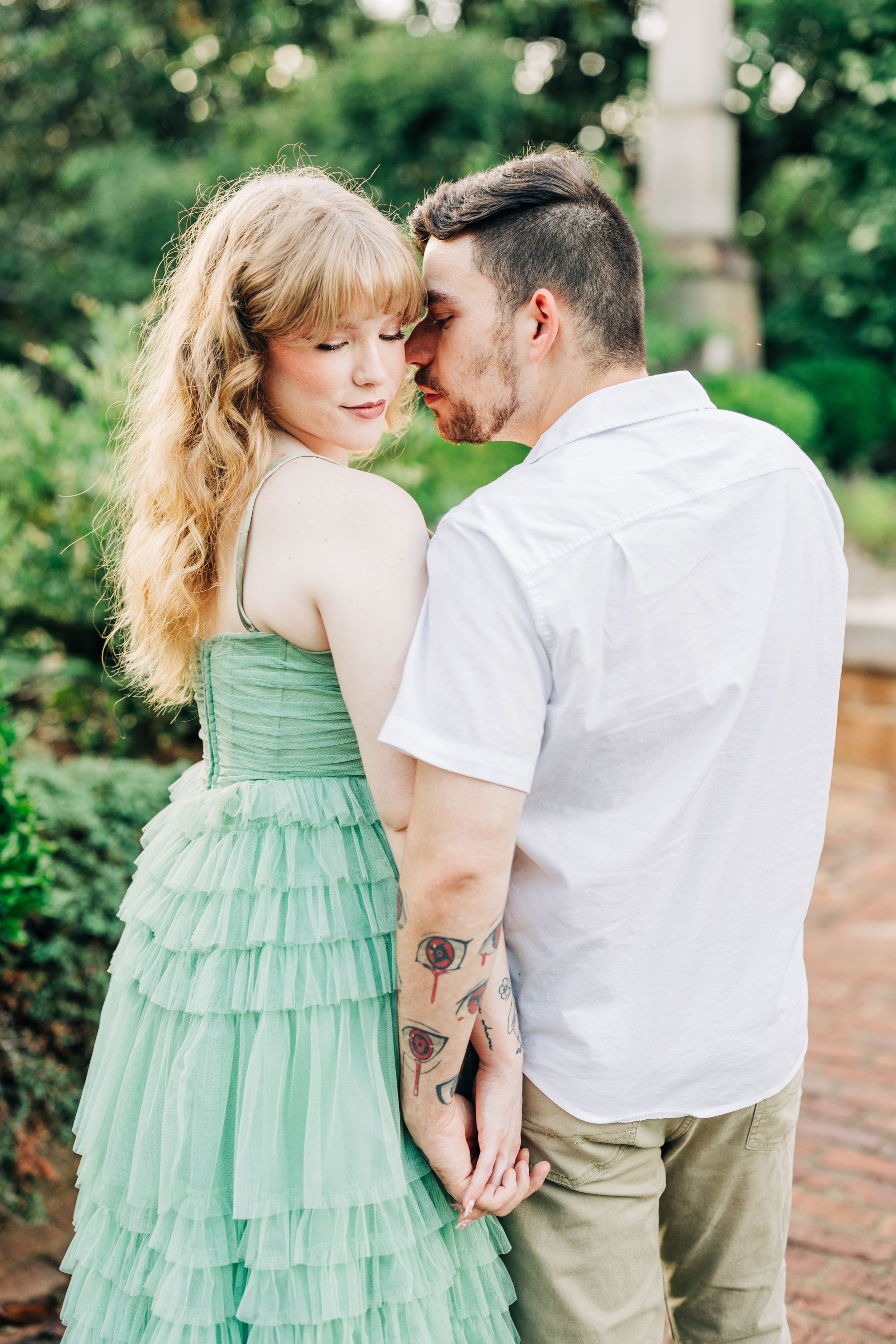 Romantic couple embraces in park setting with woman wearing mint green tiered dress and man in white shirt.