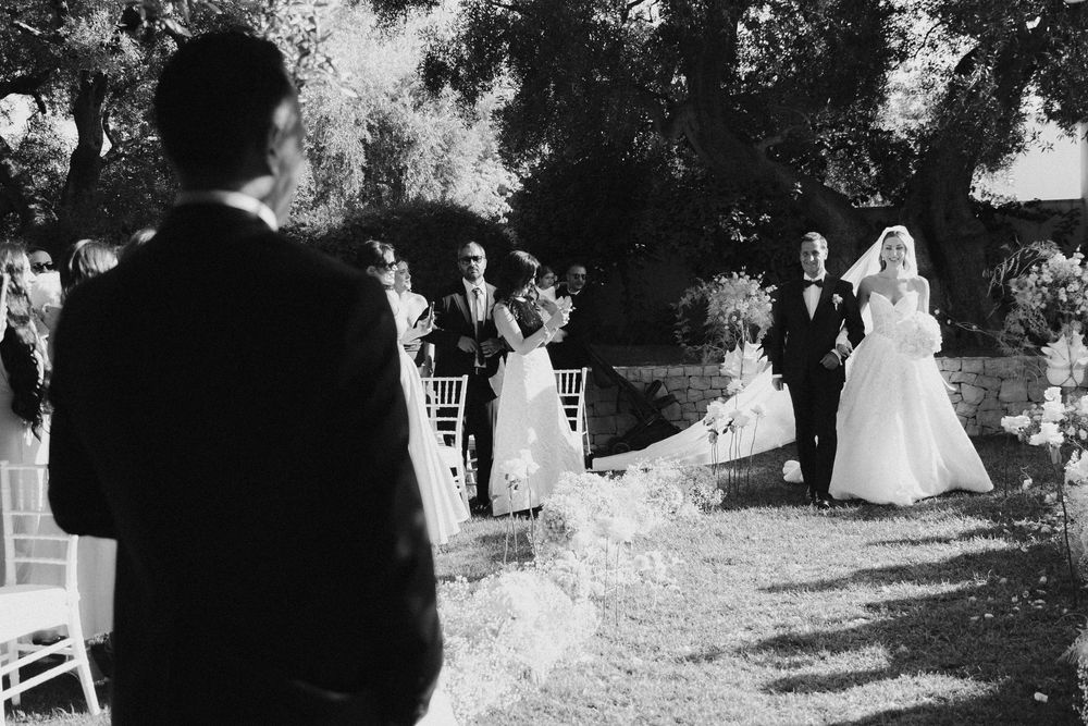 Black and white wedding photograph showing guests walking outdoors among trees during an elegant garden ceremony.