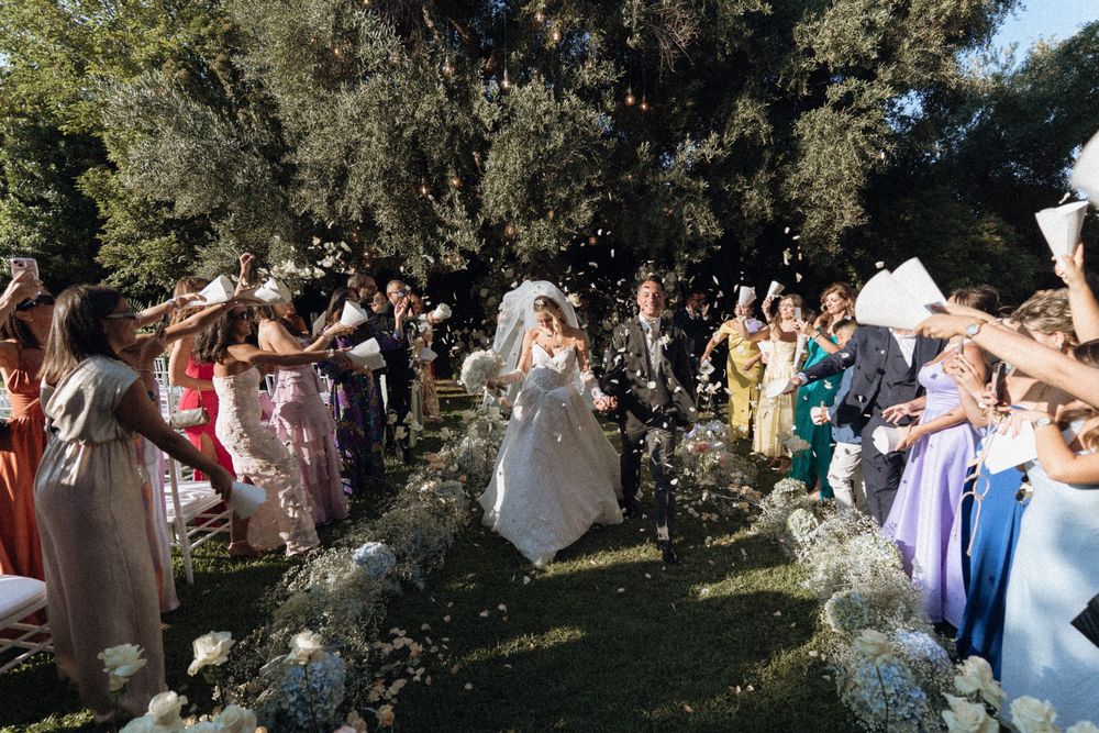 A joyful wedding party tosses confetti and flower petals in celebration outdoors under sunny skies.