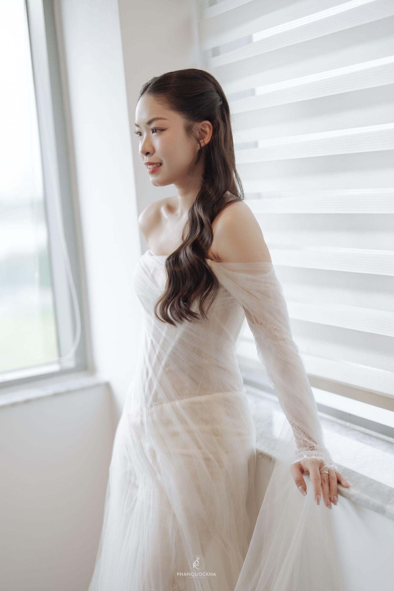 Asian bride in white off-shoulder wedding gown gazing out a window with soft natural light.