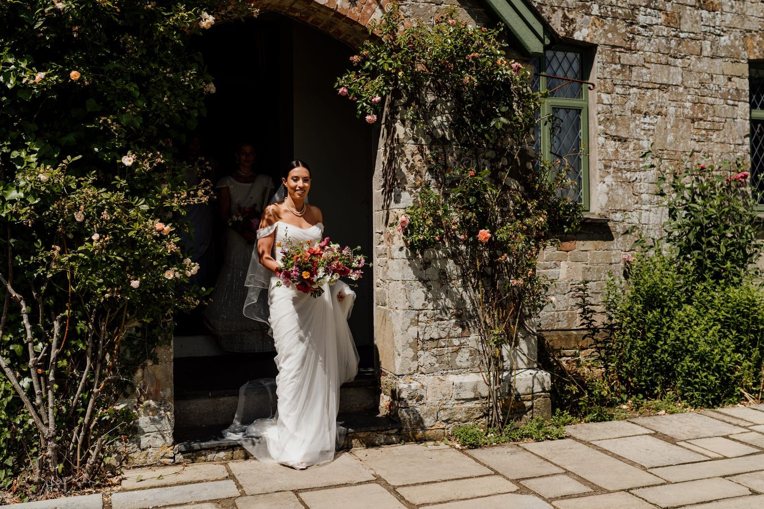 A person in a white wedding dress holds a bouquet while standing in front of a rustic stone building.