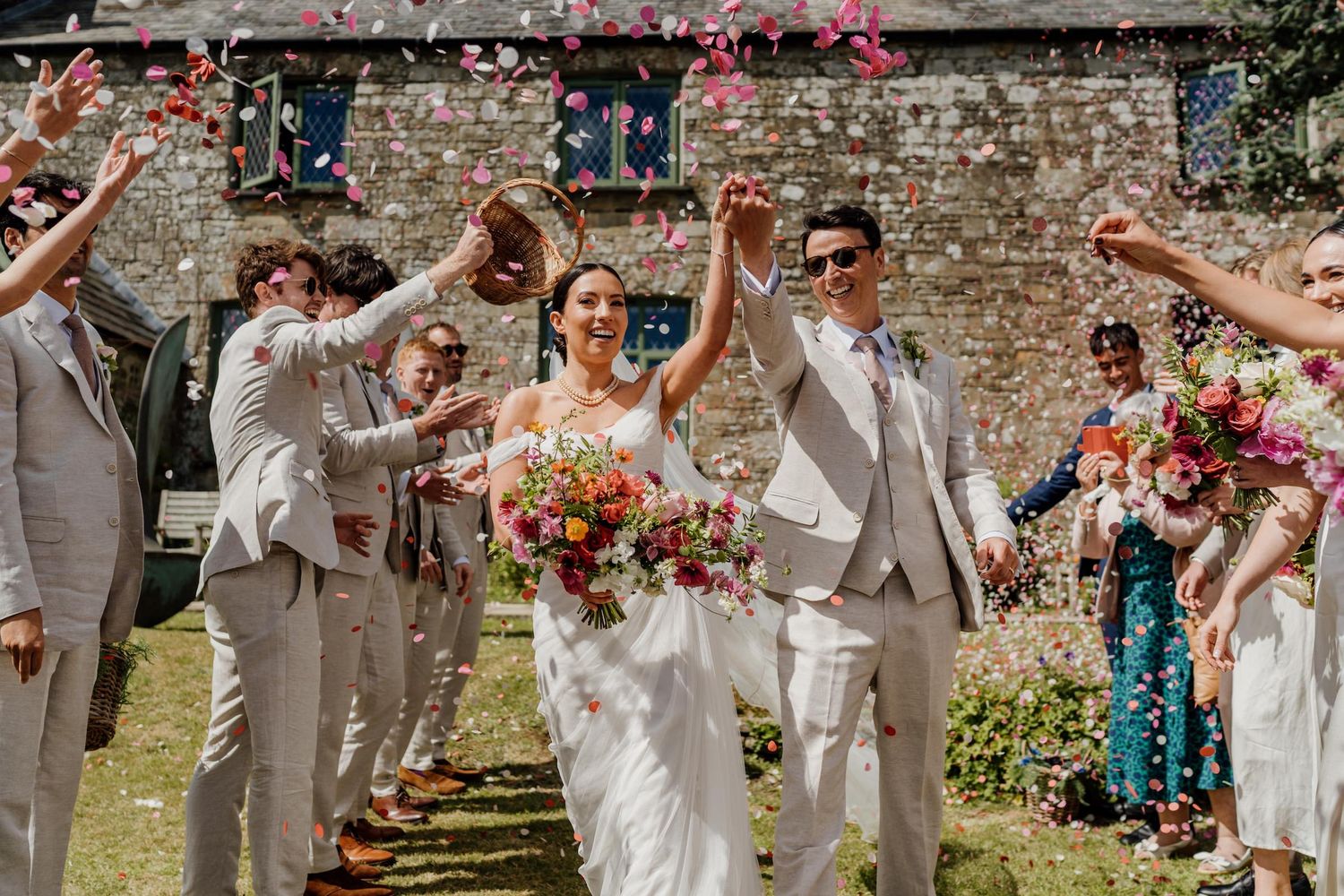 Wedding guests shower happy newlyweds with colorful flower petals as they walk down a stone-lined path in celebration.
