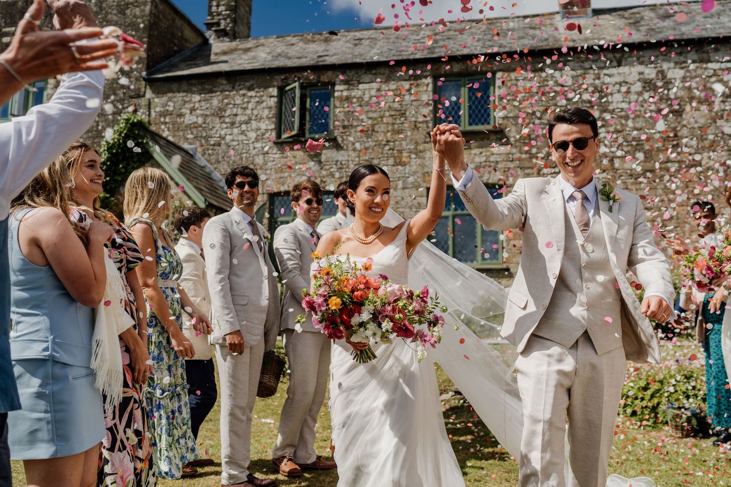 Wedding party celebrating with confetti toss outside stone wall building on sunny summer day.