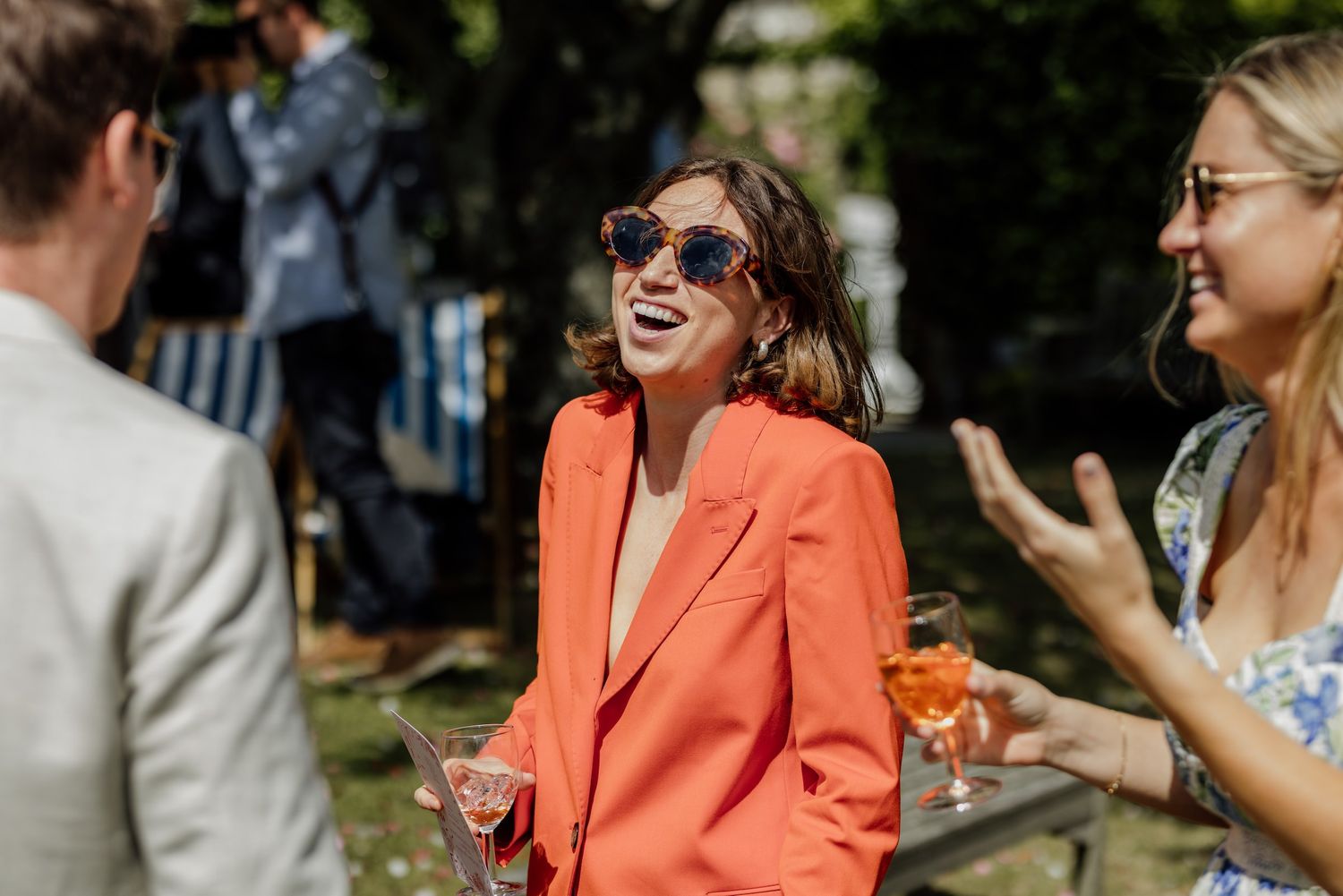 Summer wedding guest in vibrant coral blazer laughing outdoors in sunlight.