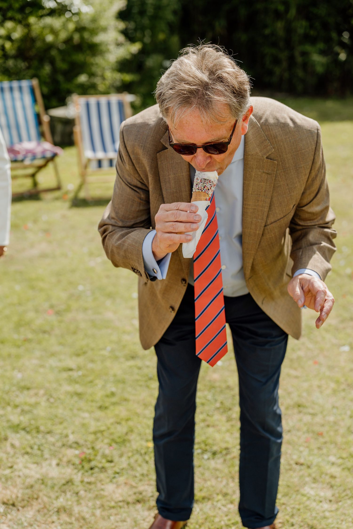 Wedding guest in brown blazer enjoying ice cream cone on sunny garden lawn.