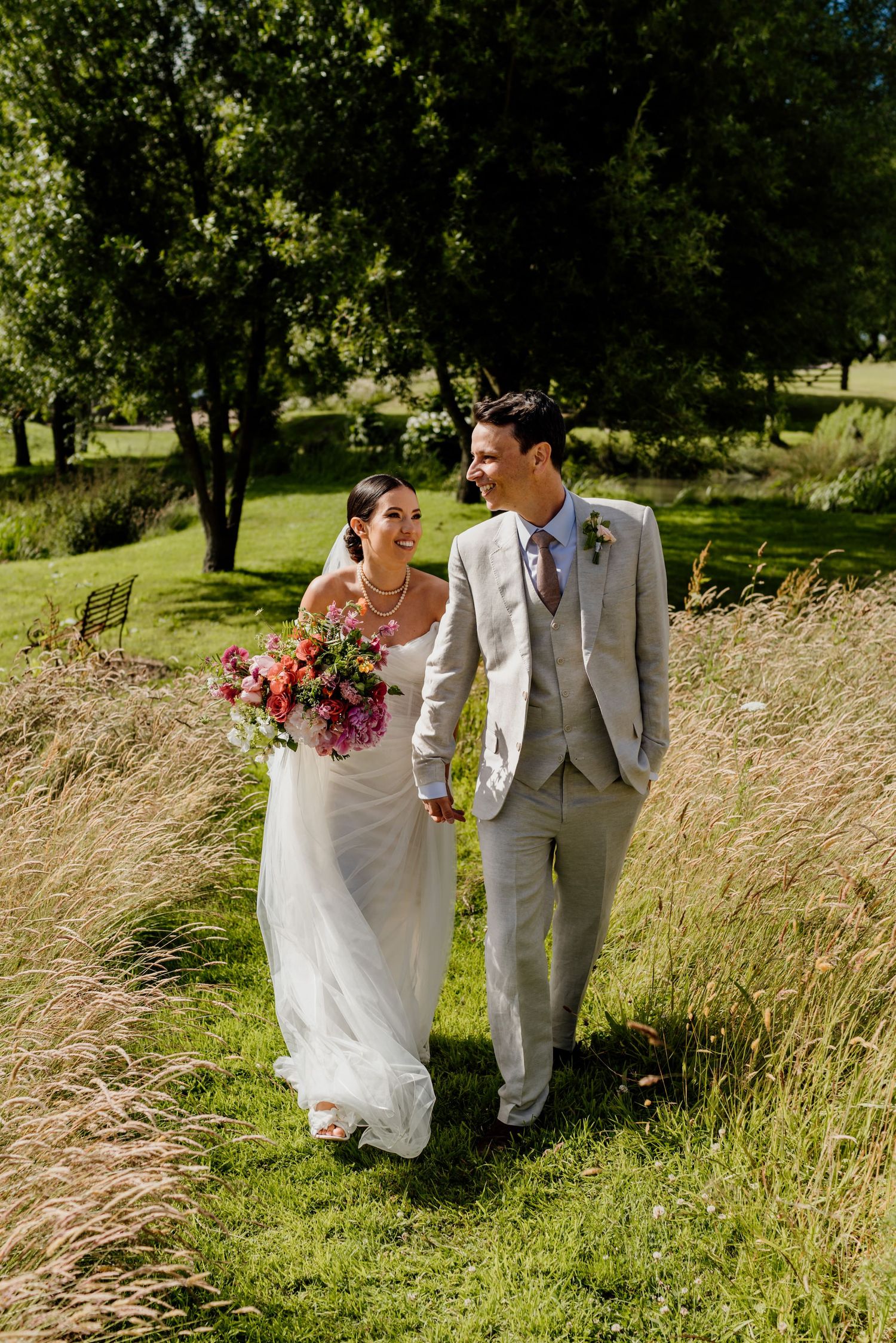 Couple walks through tall grass in a sun-lit field during their summer wedding photoshoot.