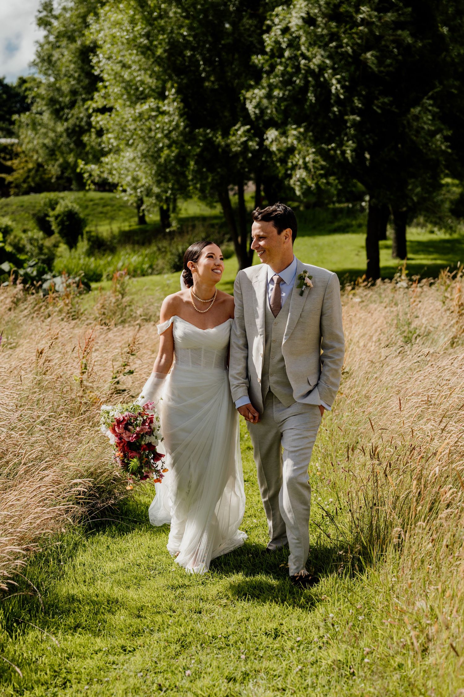 A couple walking together through tall golden grass on their wedding day in a sunlit meadow.