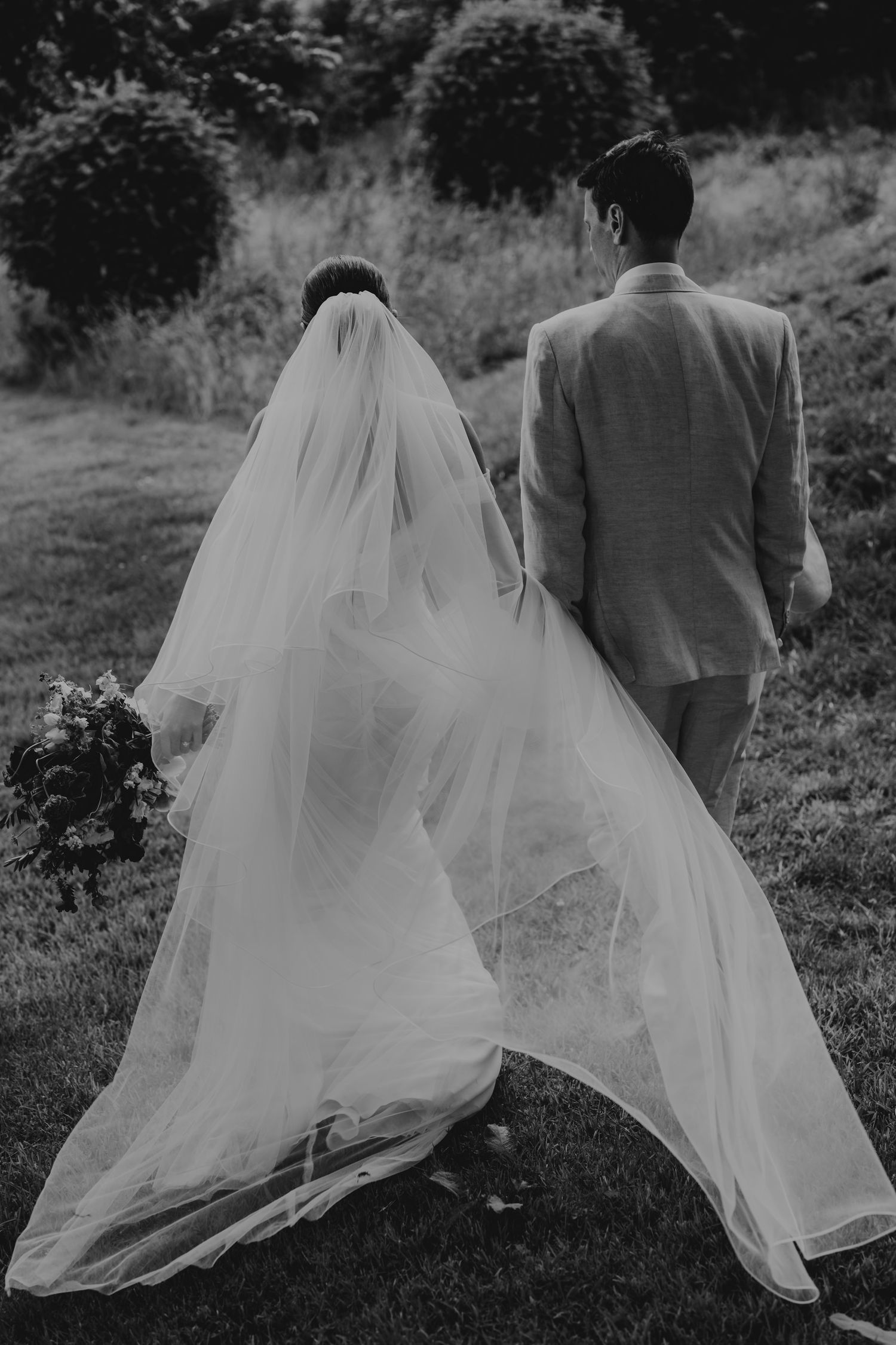 Black and white photo of newlyweds walking outdoors with a flowing white veil trailing behind on a grassy path.