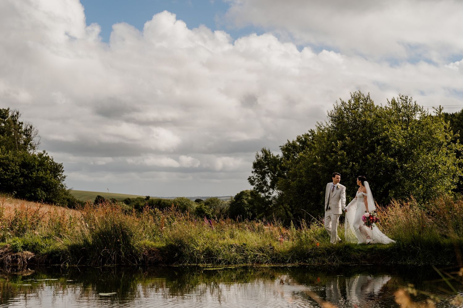 A romantic wedding scene by a pond reflecting sky and trees with dramatic cloudy backdrop.