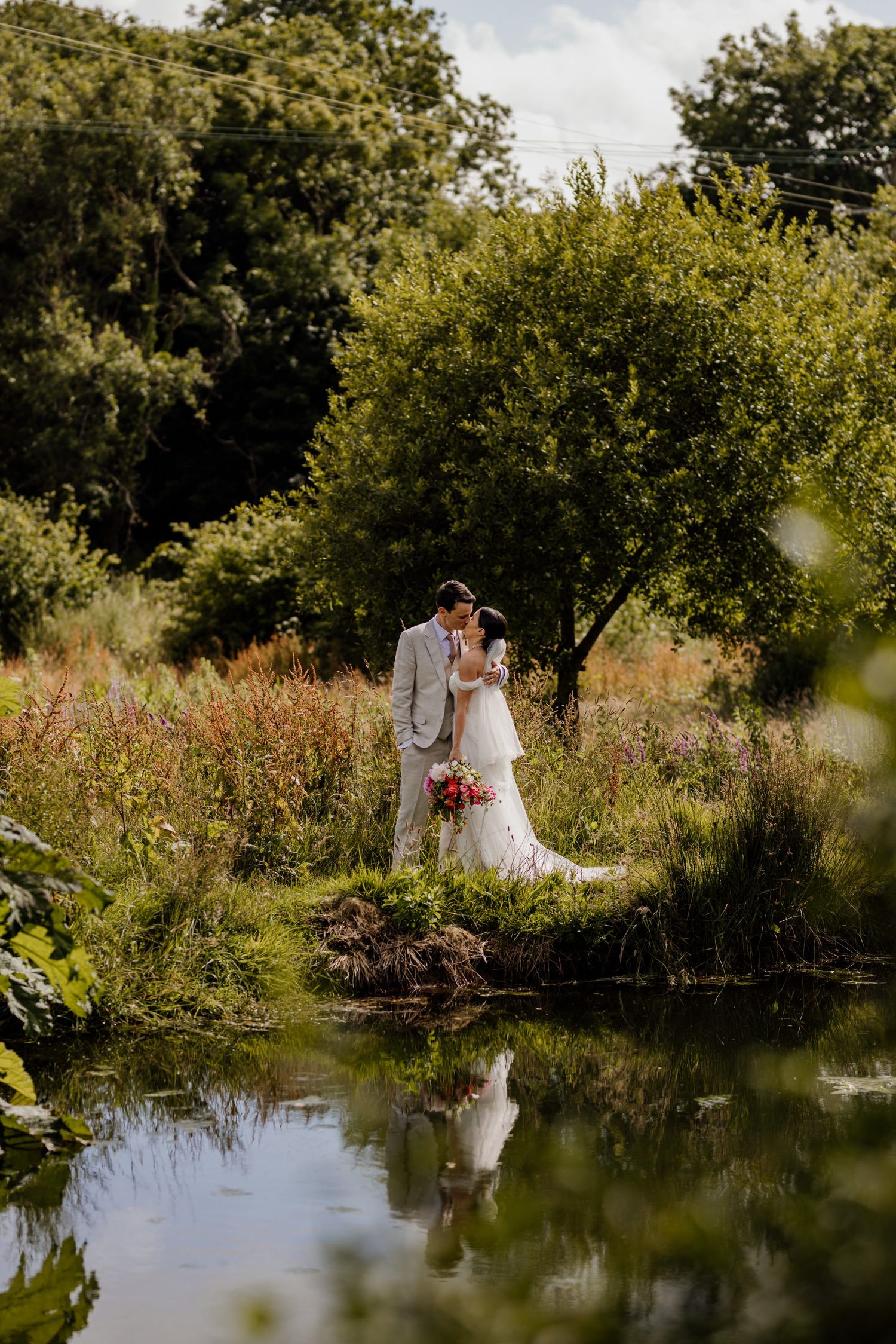 A couple in wedding attire stands together by a tranquil pond surrounded by lush summer foliage and wildflowers.