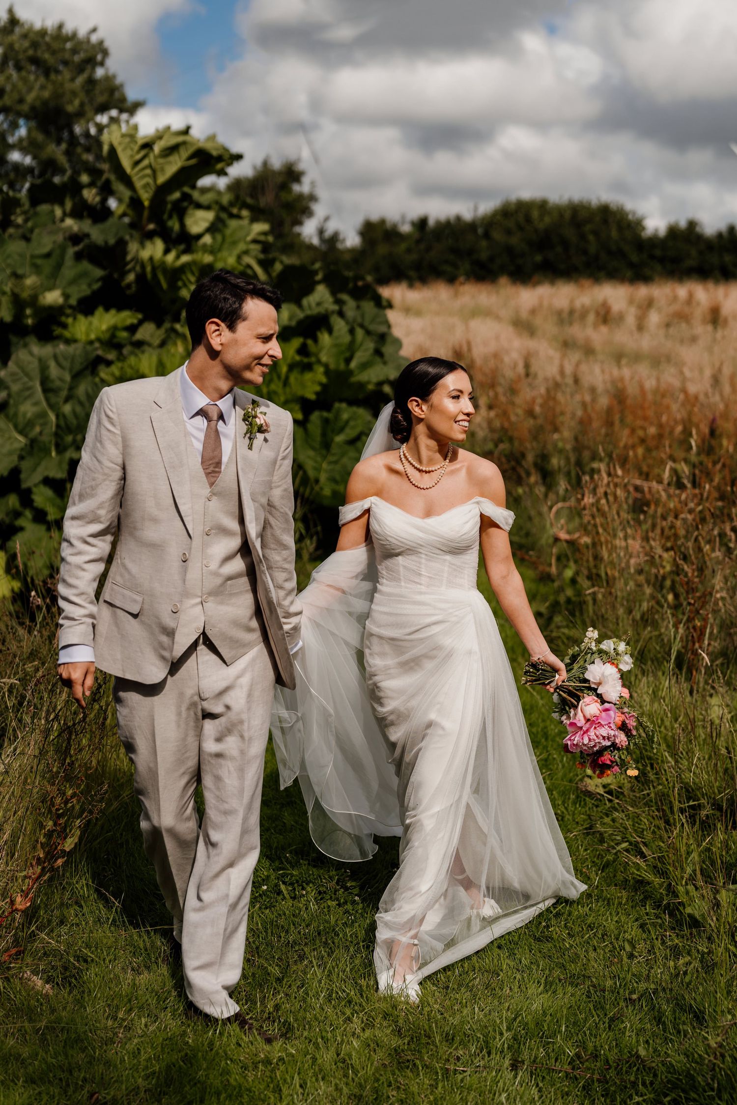 Bride in white dress holding pink bouquet walks with groom in beige suit through rustic outdoor field.