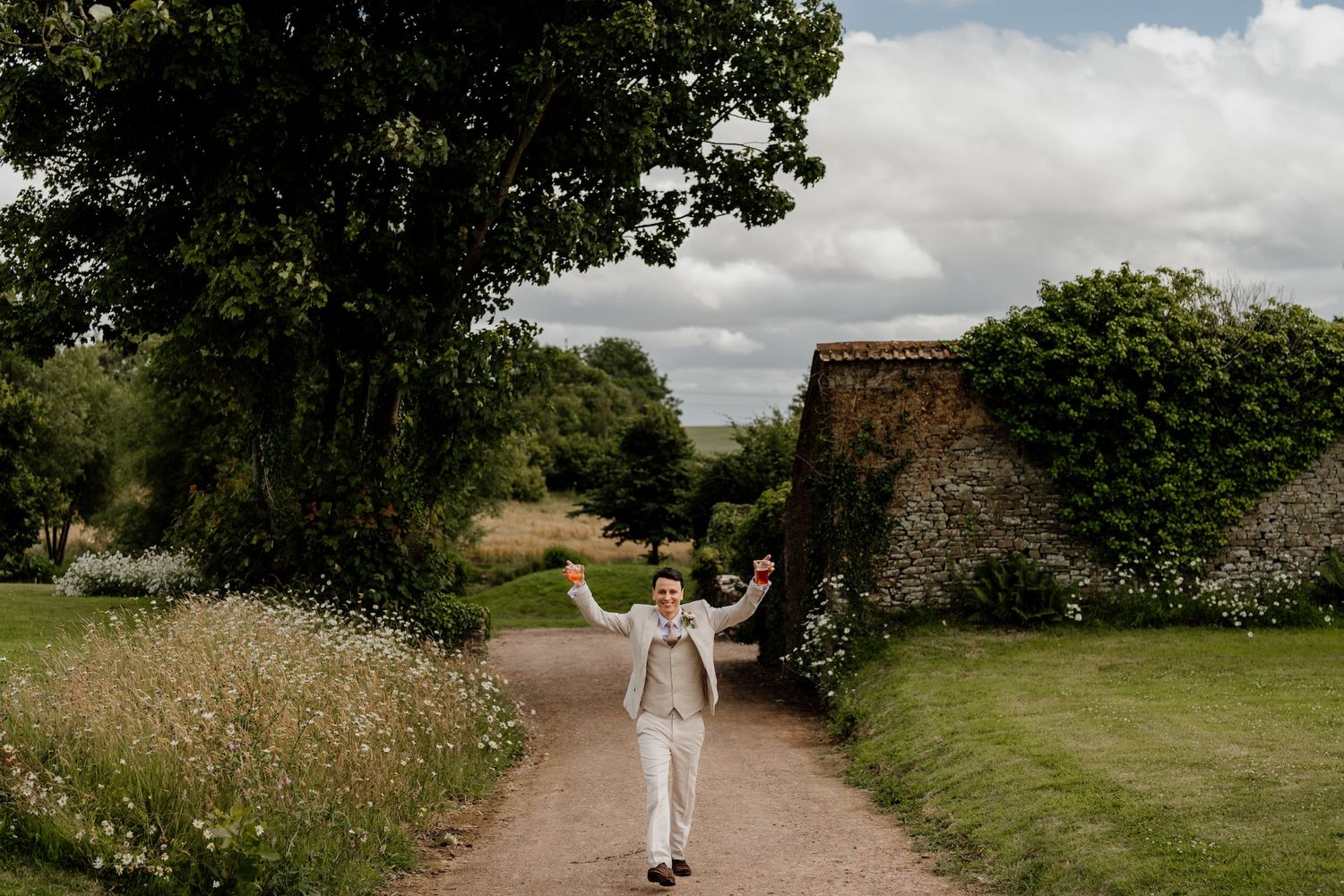 A couple walks hand in hand down a scenic countryside path surrounded by lush green trees and wildflowers.