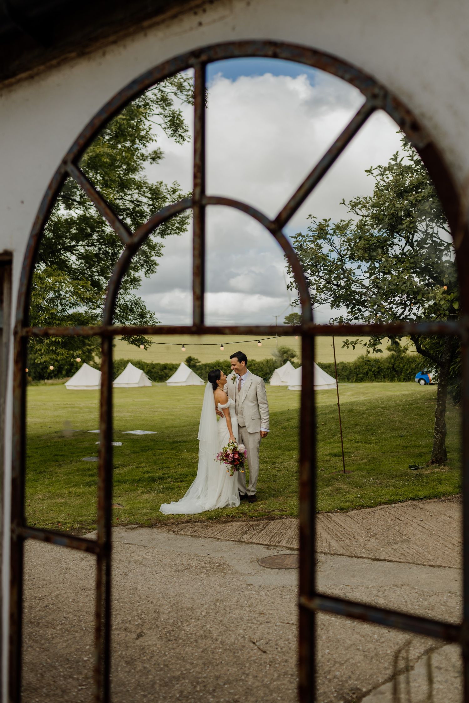 Panoramic view through arched windows of a wedding couple posing in a grassy field with white tents.