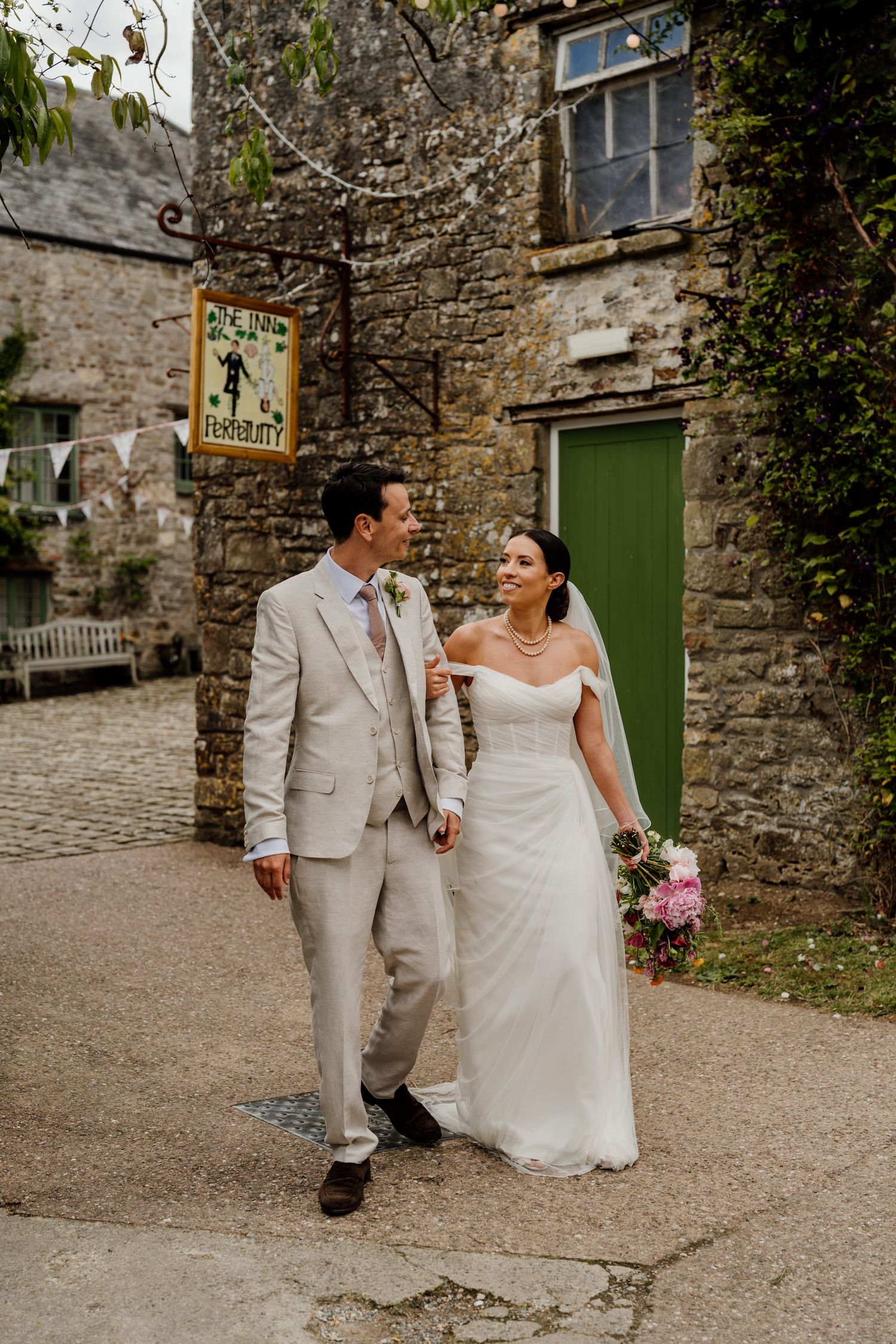 A couple strolls along a charming stone path outside a rustic building with green doors.