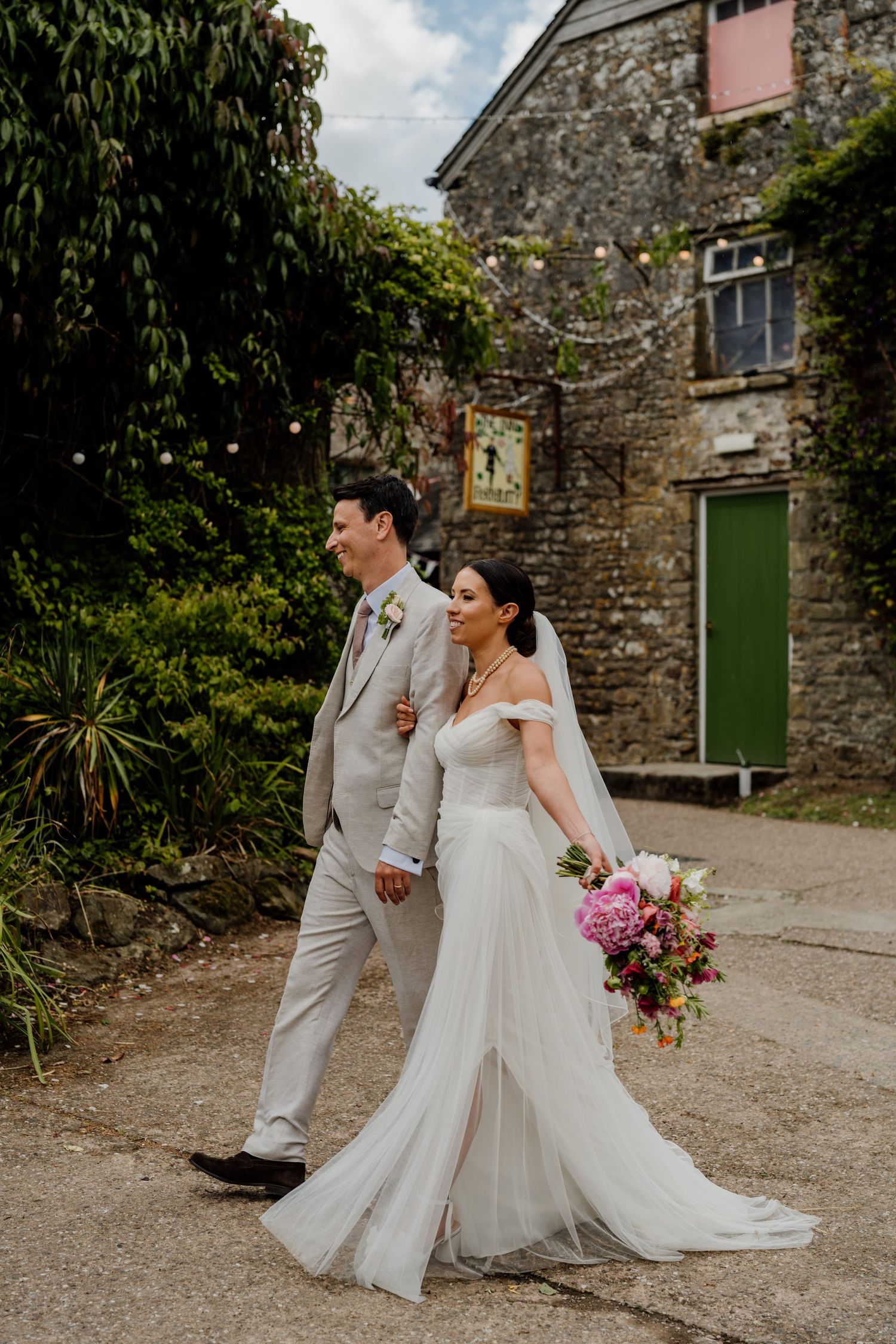 Newlyweds share tender moments outside a rustic stone building with bright pink florals.