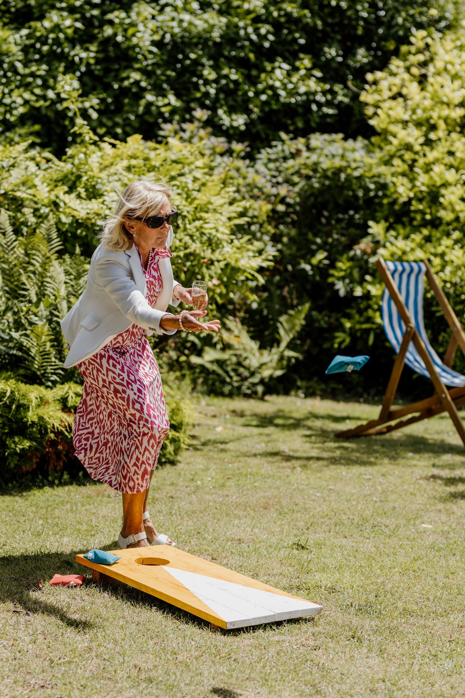 Person playing cornhole game in a sunny garden setting with deck chairs visible in the background.