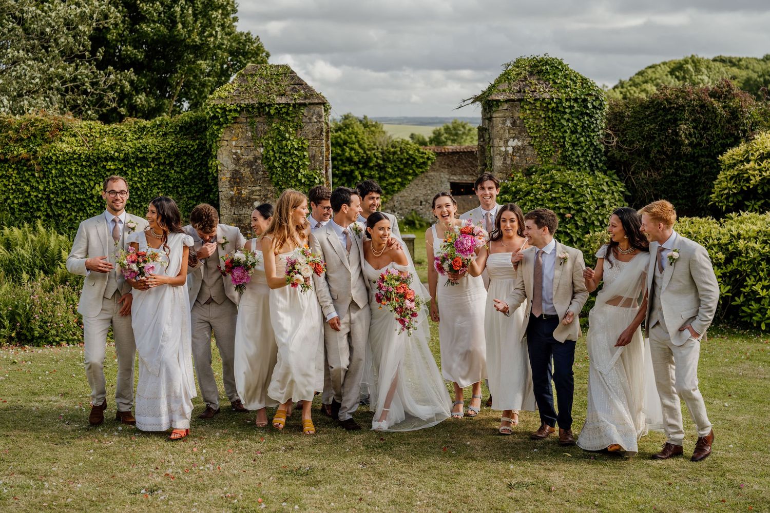 Wedding party dressed in white poses together on grassy lawn with trees and cloudy sky in background.