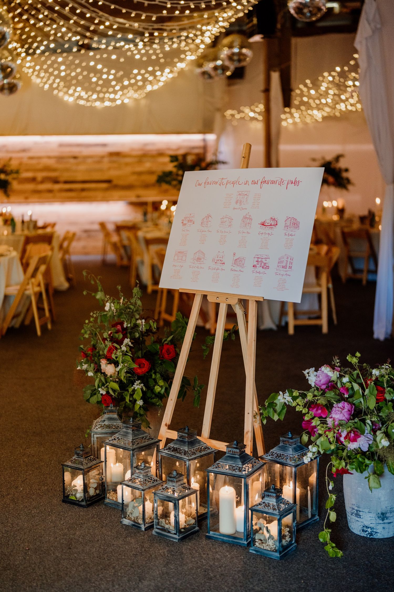 Elegant wedding reception decor with metal lanterns, string lights, and seating charts on wooden easels.