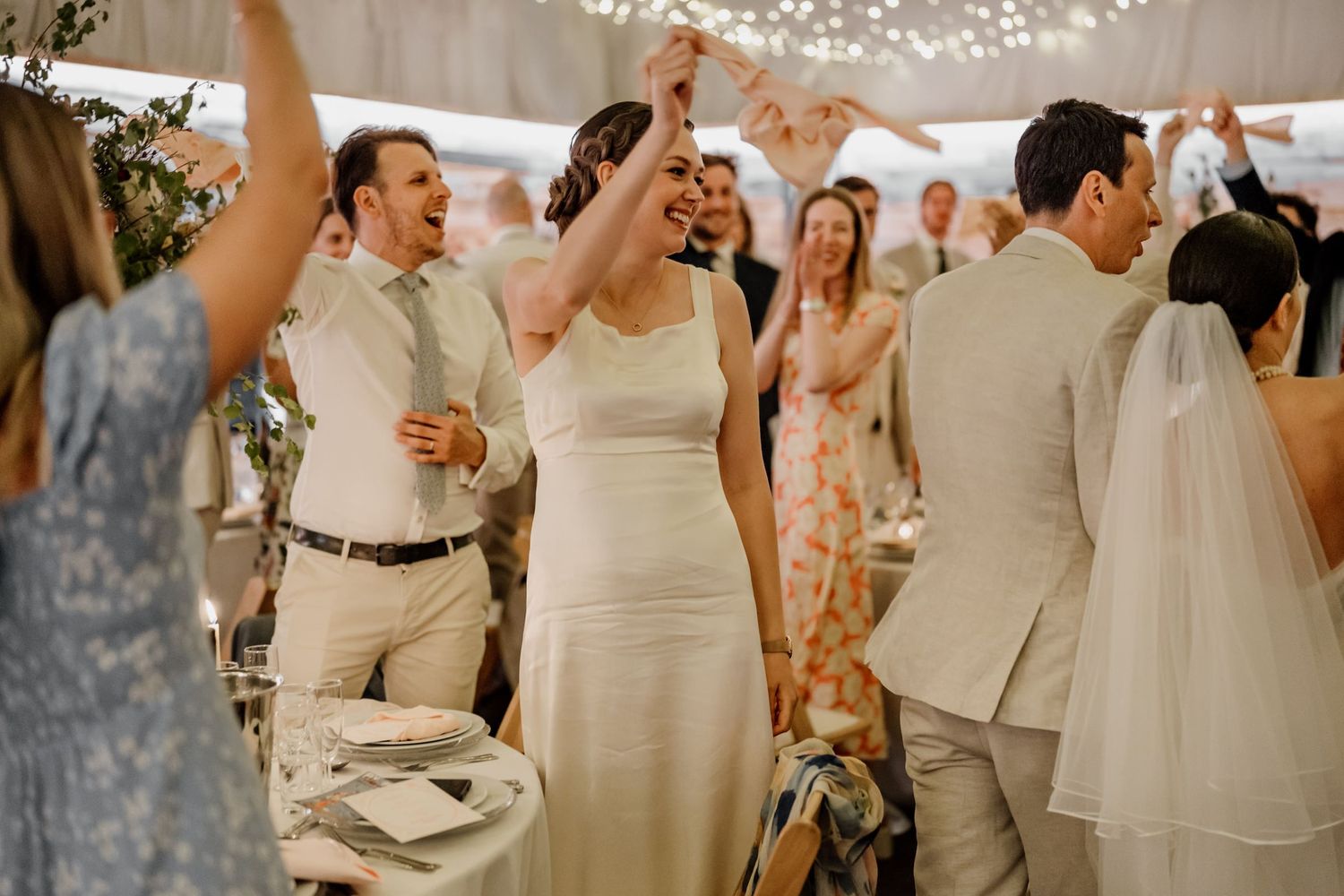 Wedding guests dancing and celebrating at a reception under string lights in an indoor venue.