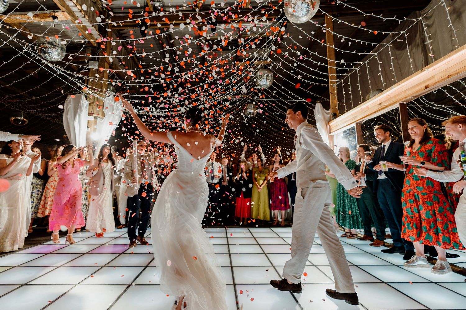 A couple dances joyfully on a lit-up dance floor under twinkling lights at their wedding reception while guests watch on.