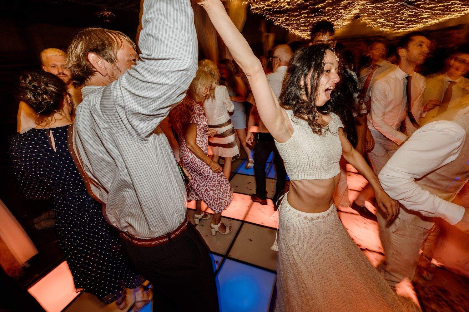 Wedding guests dancing energetically on a crowded dance floor under decorative lighting.