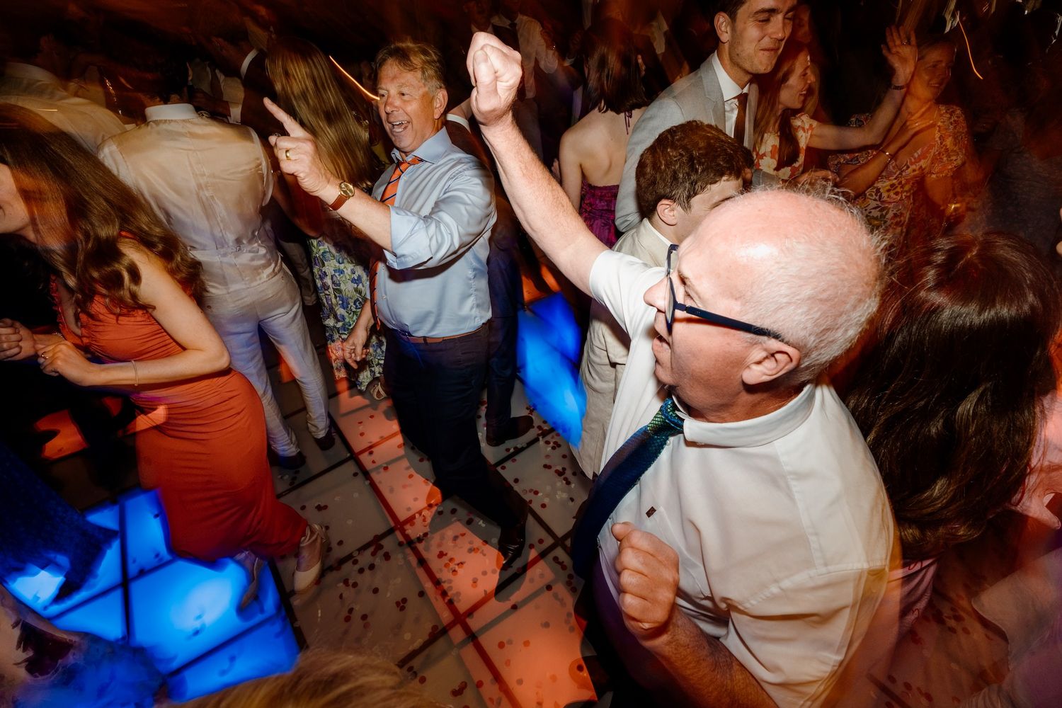 Wedding guests dancing and celebrating on a dimly lit dance floor at an indoor reception party.