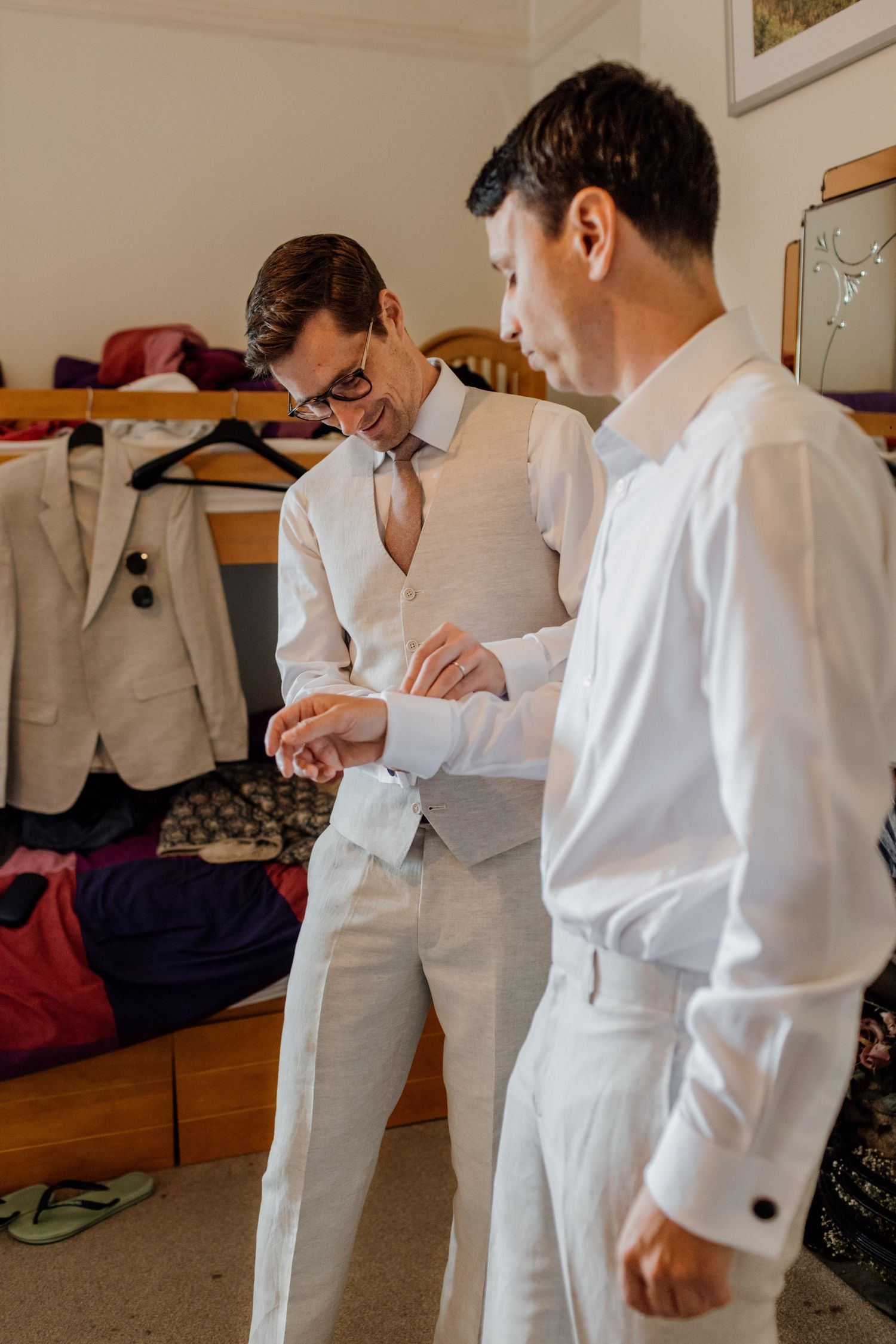 Two people in white formal attire getting dressed for an event while looking at clothing details in a room.