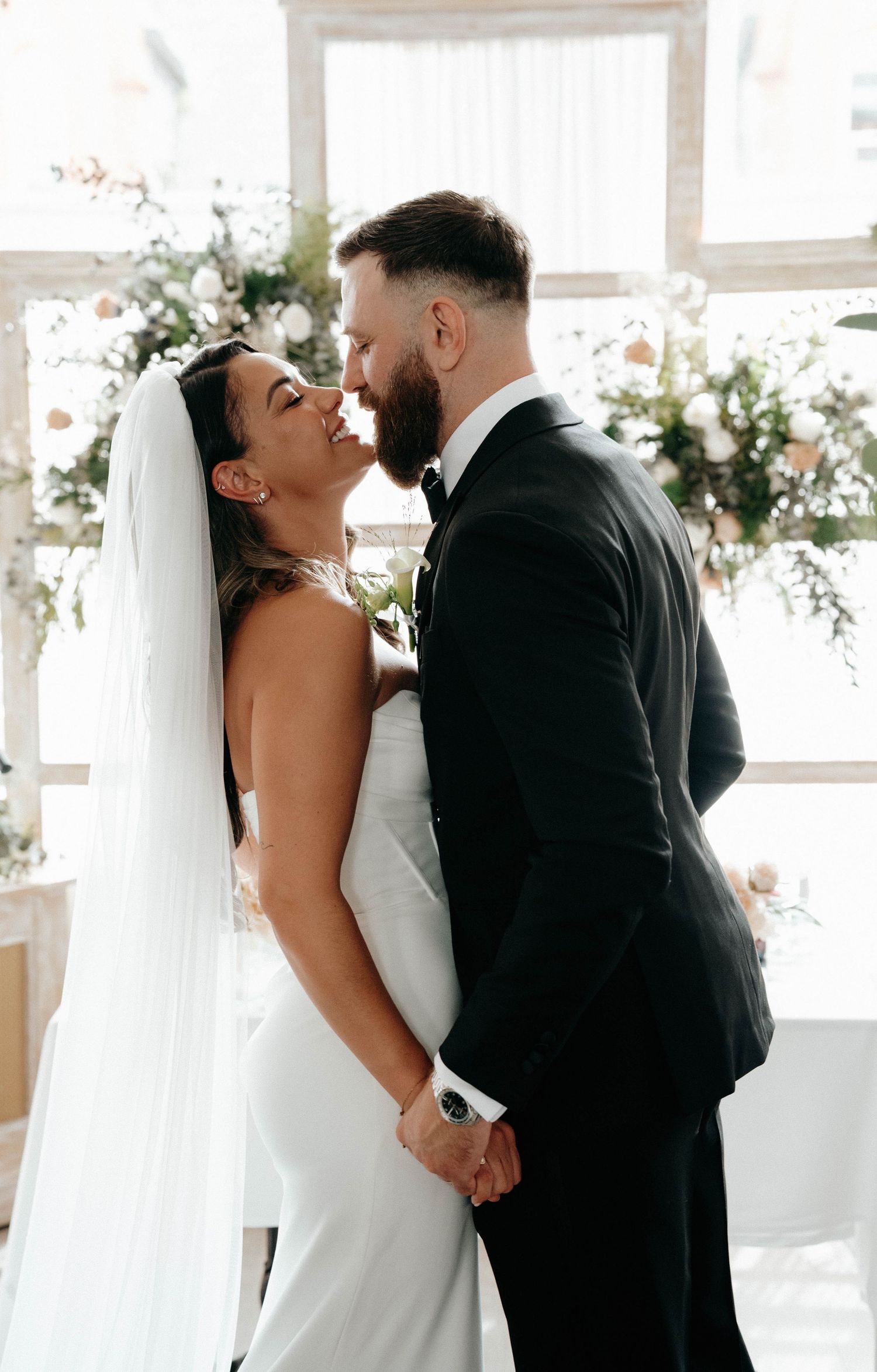 An intimate wedding moment shows a bride and groom sharing a tender kiss in a bright, white-decorated venue.