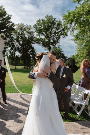 Outdoor wedding ceremony celebration with guests watching couple's first kiss under green trees.