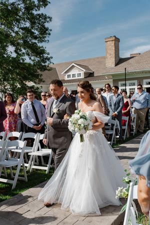 Wedding guests seated in white chairs watch outdoor ceremony with bridesmaids in light blue dresses at elegant venue.