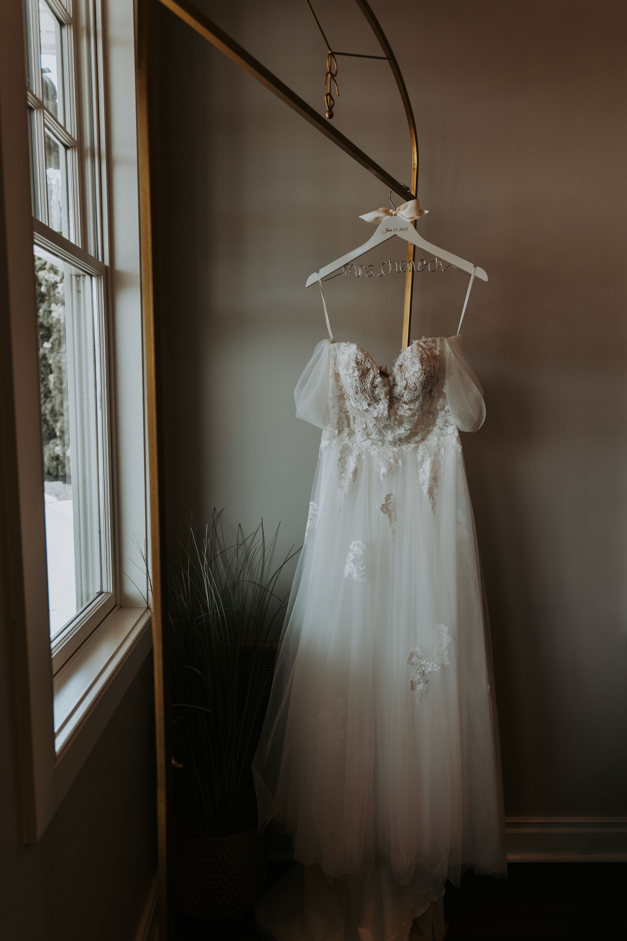 White lace wedding dress hangs elegantly by a window, showcasing delicate details and flowing fabric.