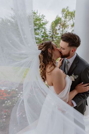 Romantic first look moment with flowing veil in front of white columns and blooming flowers.