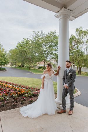 Wedding couple and bridal party posing under white columns with flower gardens in background.