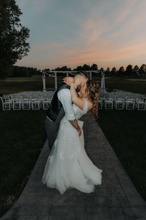 Romantic sunset wedding ceremony setup with white chairs and couple embracing on stone pathway.