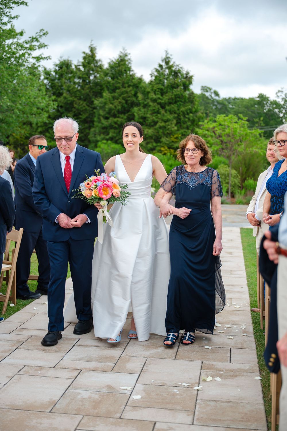 A bride is escorted by her parents down the outdoor stone aisle at the Lakehouse in Halifax, MA