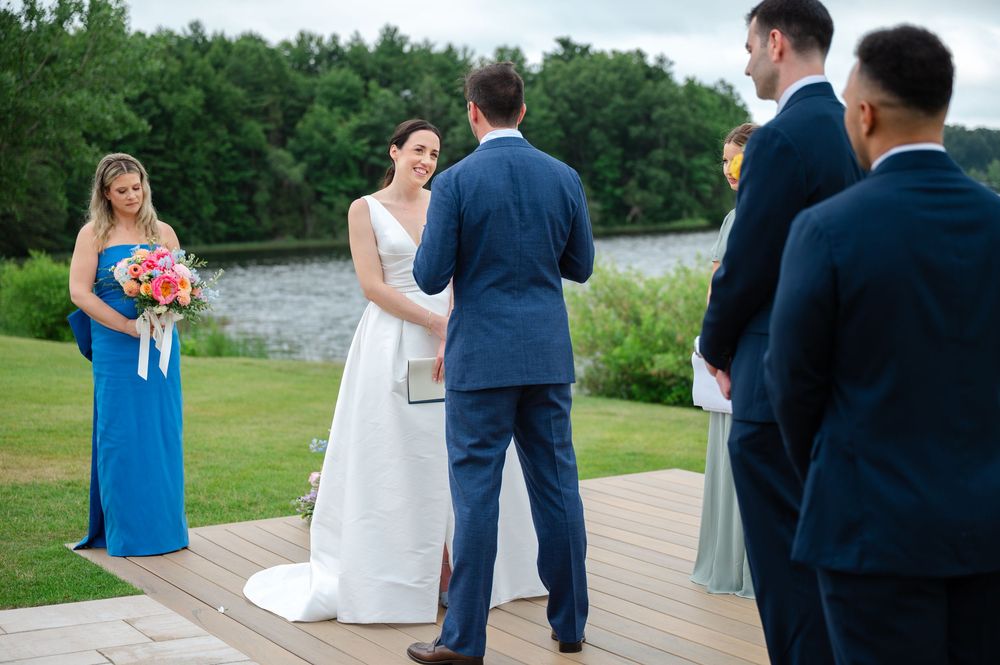 A couple exchanges wedding vows during an outdoor ceremony in a lush green field with a lake in the background.