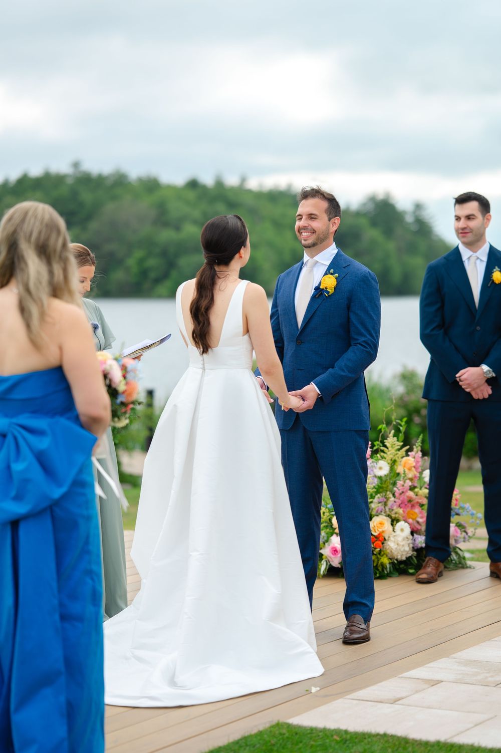 Wedding party in blue attire stands on waterfront deck during outdoor ceremony with white floral arrangements.