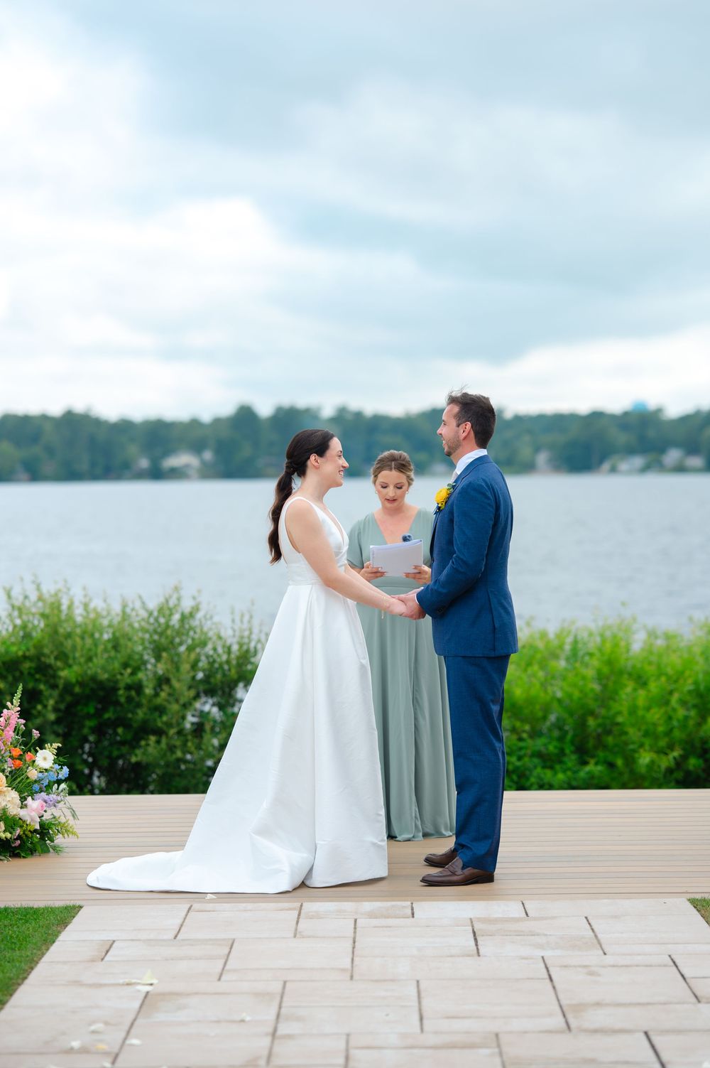 A bride and groom share a tender moment during their lakeside wedding ceremony on a stone patio.