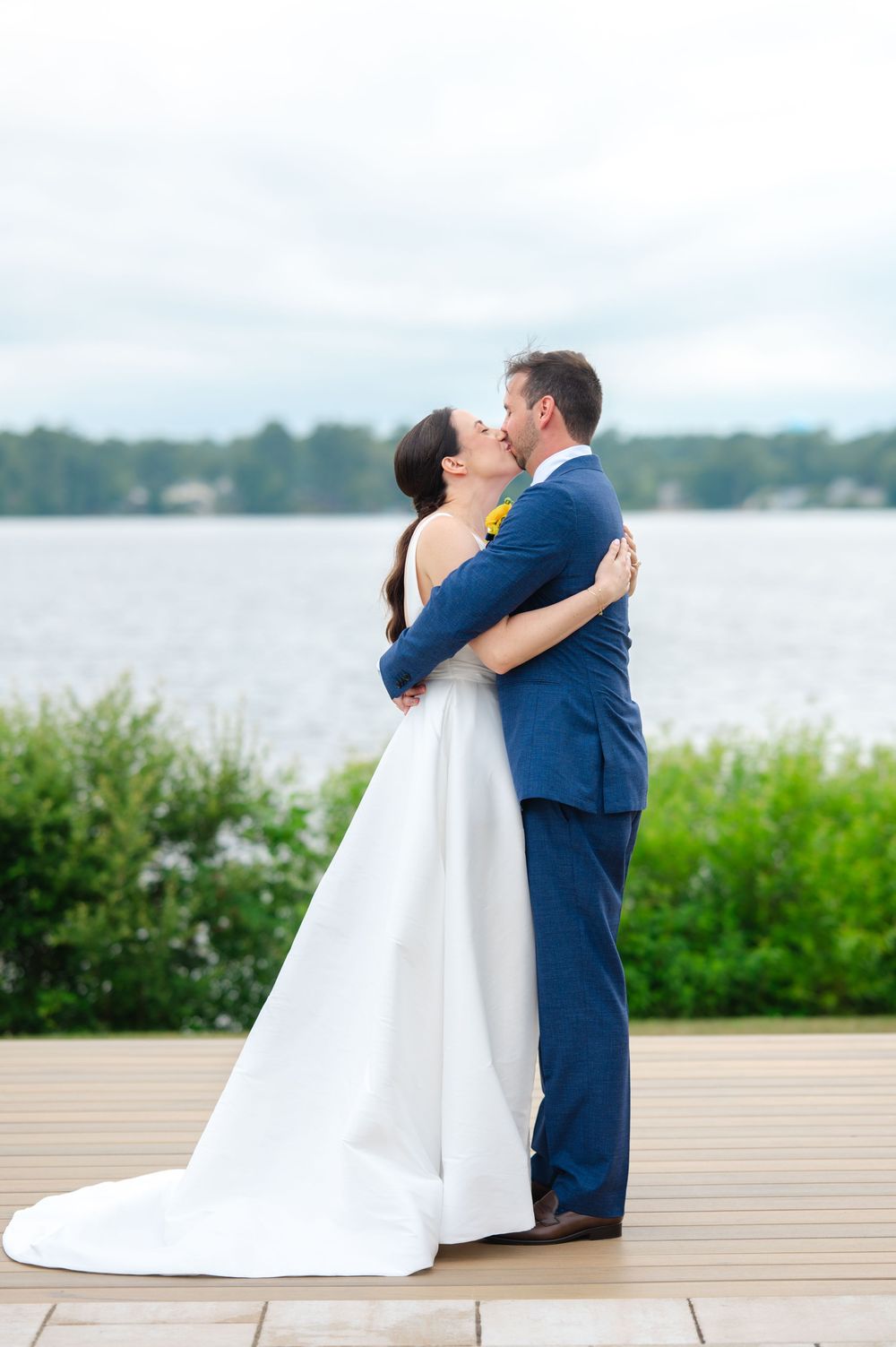 A couple in wedding attire embrace on a wooden deck overlooking a serene lake with greenery in the background.