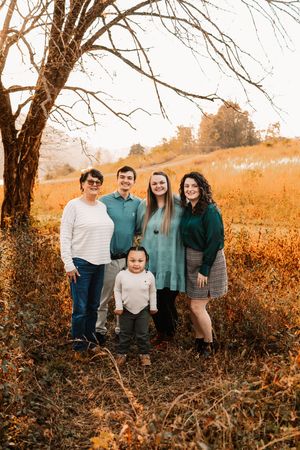 A multigenerational family of five poses together in a golden autumn field beneath a bare tree at sunset.