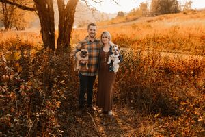 Happy couple holding two small dogs in golden autumn field during sunset photography session.