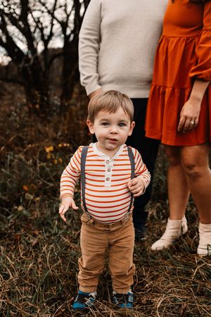 An adorable toddler in striped shirt and suspenders stands in front of his parents in an autumn setting.