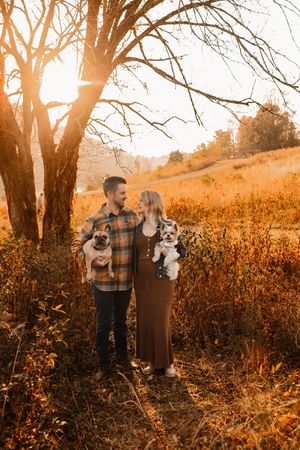 Couple with two dogs standing under bare tree in glowing golden hour autumn landscape.