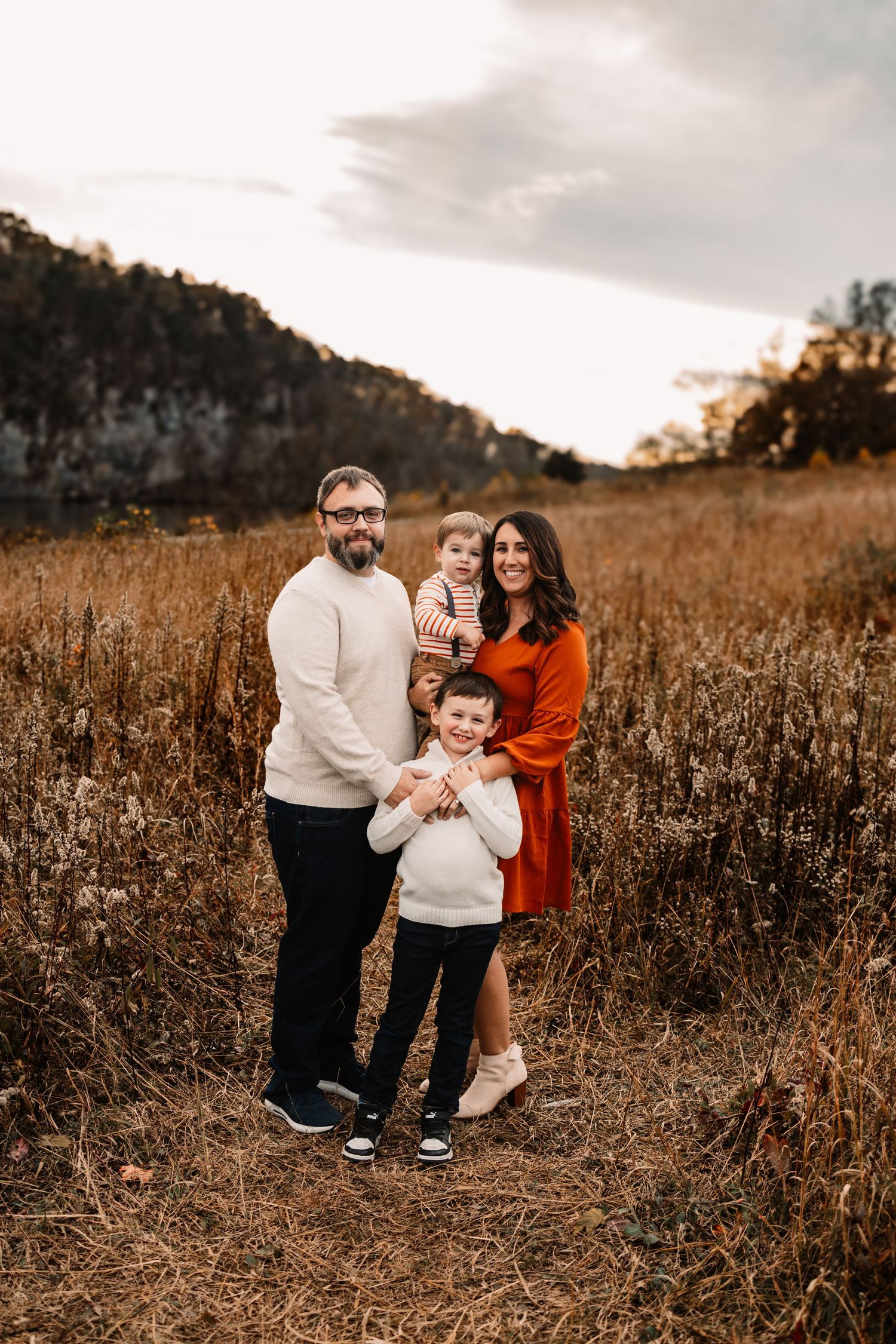 A smiling family of four poses together in a golden autumn field with rocky bluffs in the background.