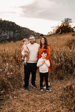 A happy family of four stands together in a dry autumn meadow with a rocky hillside behind them.