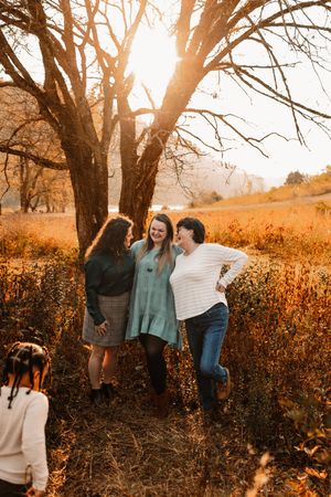 Three women laugh and embrace near a bare tree in a sunlit autumn meadow during golden hour.