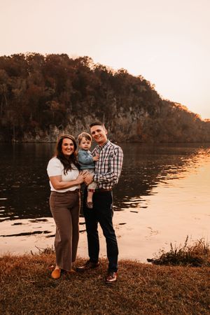 Smiling family of three posing by reflective lake with colorful autumn cliffs at sunset.